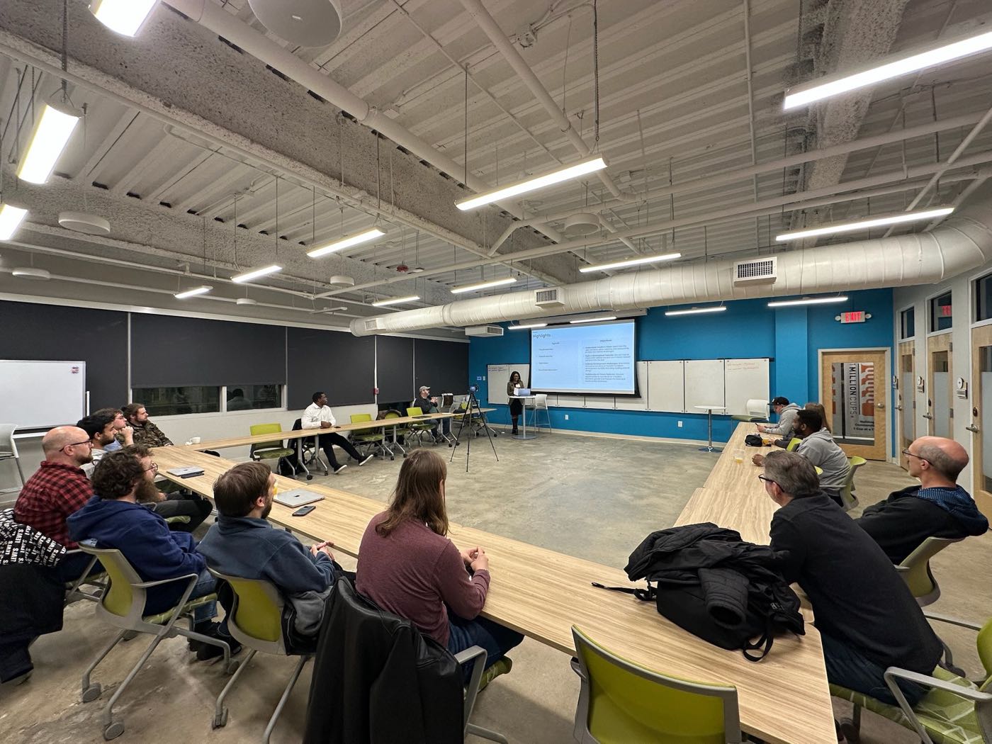 An evening meetup at Flywheel Coworking in downtown Concord: around fifteen people seated at long tables, watching a speaker present slides.