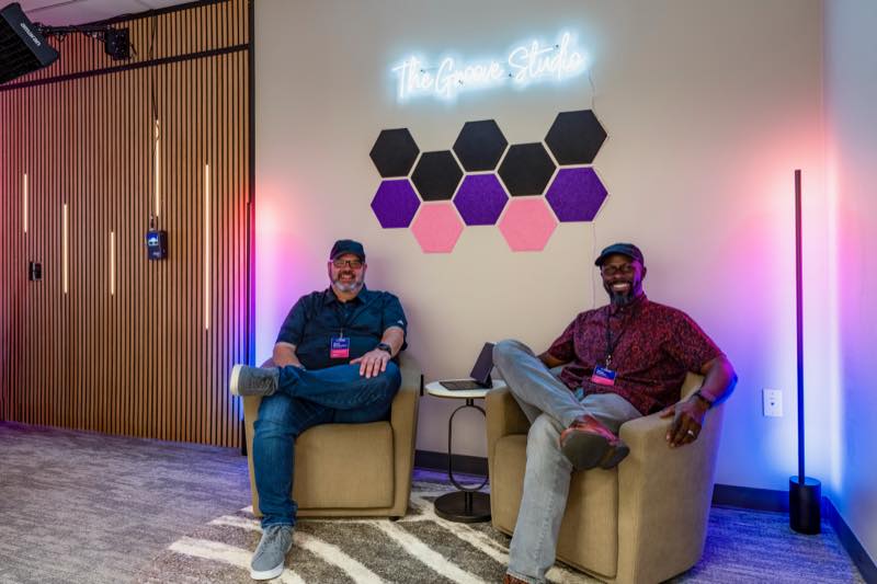 Mark Shropshire and Bayo Fodeke sitting in The Groove Studio's lounge, beneath the studio's neon sign and a wall of hexagon acoustic panels.