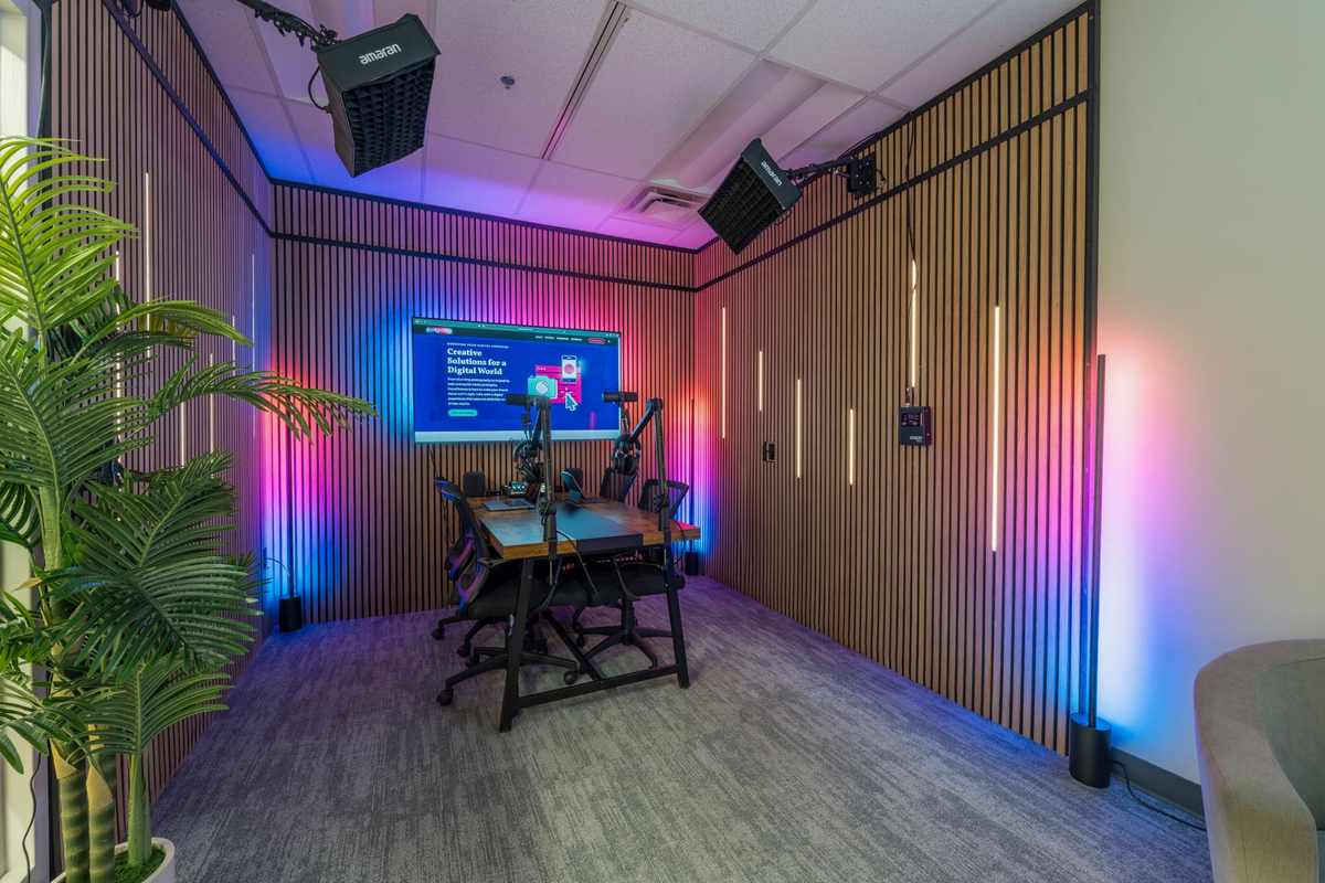 The Concord studio: a podcast table with microphones on boom arms and four chairs, facing a wall-mounted screen, with colored LED light washing the slatted wood walls.