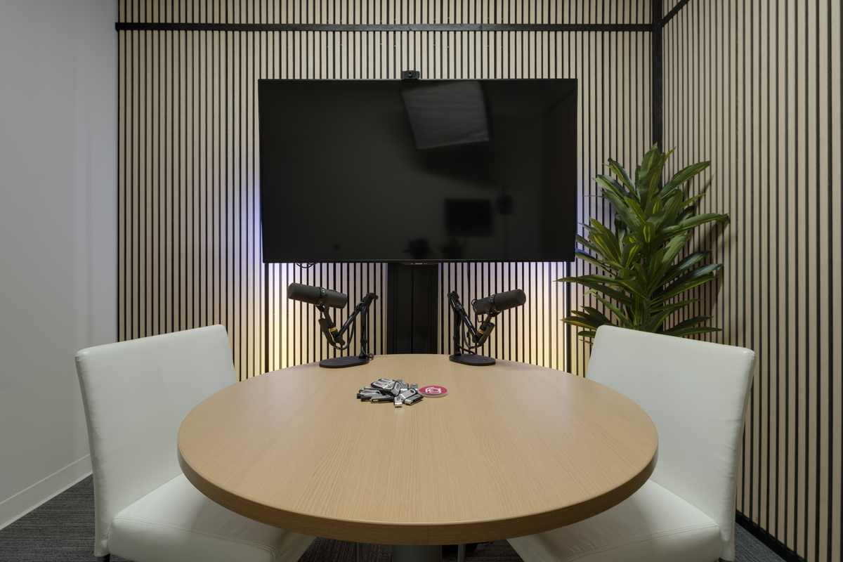 The Winston-Salem studio: a round table with two microphones on boom arms and two chairs, facing a wall-mounted screen against slatted wood walls.